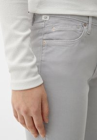 Jeans de mezclilla gris claro con textura sutil, que cuentan con un diseño de cinco bolsillos, botones de tono cobre y una etiqueta blanca de la marca en la cinturilla.