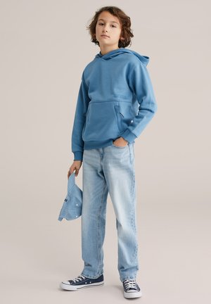 Sinine kapuutsiga dressipluus esitaskuga, kombineeritud helesiniste teksapükstega. Laps hoiab kätte sobiva sinise nokamütsi ning kannab tumesiniseid tenniseid.