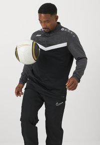 Schwarzes und graues Sportanzug mit einem gemusterten Oberteil, das ein weißes Chevron-Design und Logo-Details aufweist. Hält einen schwarz-weißen Fußball.
