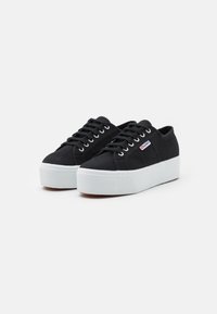 Schwarze Canvas-Sneaker mit einer erhöhten weißen Gummi-Sohle, ausgestattet mit runden Schnürsenkeln, silbernen Ösen und einem kleinen Logo an der Seite.