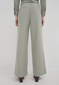 Pantalons gris clair à jambes droites avec poches passepoilées au dos, portés avec des chaussures à talons bruns, vus de dos.