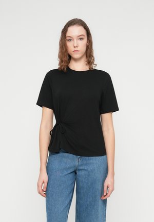 Sort T-shirt med korte ærmer og knudedetalje på venstre side, lavet af blødt stof. Kombineret med lyseblå jeans med vide ben.