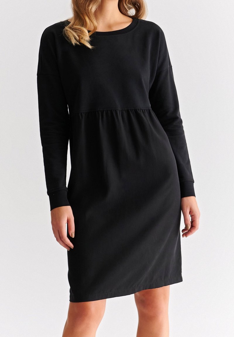 Femme portant une robe noire à manches longues, longueur genou, avec une taille froncée, debout devant un fond blanc uni.