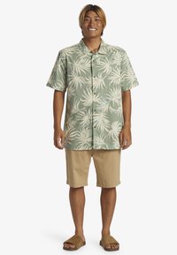 Camisa de manga corta abotonada en verde claro con un patrón de hojas de palma en blanco y azul, combinada con pantalones cortos beige y sandalias de slide marrones.