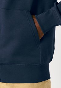 Sweat à capuche bleu marine avec poignets et taille côtelés, doté d'une poche avant et d'une texture lisse, partiellement visible avec un pantalon beige.