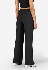 Čierne nohavice s širokými nohavicami a elastickým pásom na stiahnutie, hladká textúra a dve zadné vrecká. Detail loga na pravom vrecku.