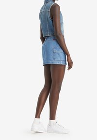 Denimväst och shorts-set i ljusblått. Västen har framfickor och är ärmlös; shortsen har en bakficka och fransade kanter. Vita sneakers.