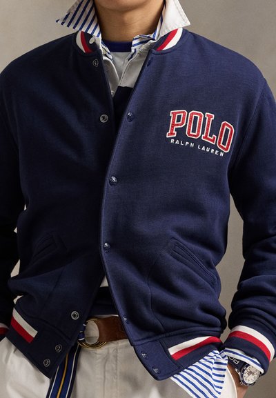 Moški, ki nosi mornarsko modro univerzitetno jakno Polo Ralph Lauren čez črtasto srajco, bež hlače in uro na levi zapestju.