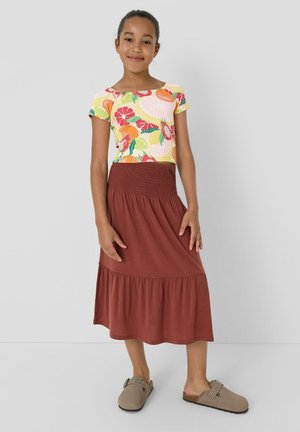 Mädchen steht da und trägt ein buntes Kurzarm-Top mit Fruchtmuster, einen rostfarbenen Midi-Rock und beigefarbene Slip-On-Schuhe vor einem schlichten Hintergrund.