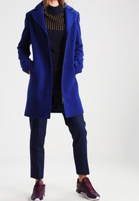 Cappotto di lana blu con colletto a rever, abbinato a un maglione scuro con dettagli dorati, pantaloni blu navy e sneaker bordeaux.