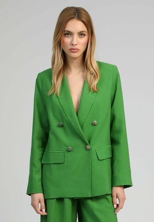 Femme aux longs cheveux blonds portant un blazer vert vif à double boutonnage et un pantalon assorti, debout devant un fond uni.