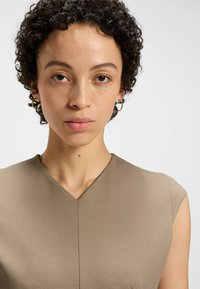 Tan sleeveless top featuring a V-neck design and smooth texture, with a subtle sheen. Model has short curly hair and simple hoop earrings.