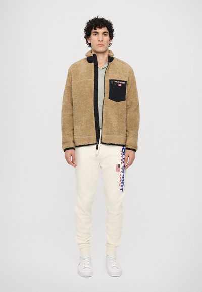 Uomo che indossa una giacca in fleece beige con tasca nera sul petto, pantaloni sportivi cream Polo Sport con logo e sneakers bianche, in piedi su uno sfondo semplice.