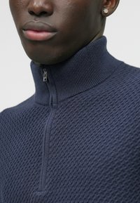 Maglione zip-up blu navy con un motivo tessuto testurizzato. Presenta un colletto alto, chiusura a mezza zip e dettagli a coste sul colletto e sull'orlo.