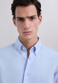 Chemise bleue claire à col pointu, avec de petits boutons jaunes et une texture en coton doux. Aucun motif ou logo visible.