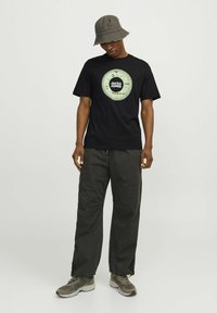 Zwarte t-shirt met een ronde grafische print in groen en zwart; gecombineerd met donkere, loszittende broeken en een grijze bucket hat. Grijze sneakers maken de outfit af.