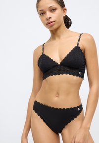 Juoda nėriniais puošta bralettė ir bikinio komplektas su banguotais kraštais bei logo dirželiais. Minkšta medžiaga su subtilia tekstūra ir prigludusiu dizainu.