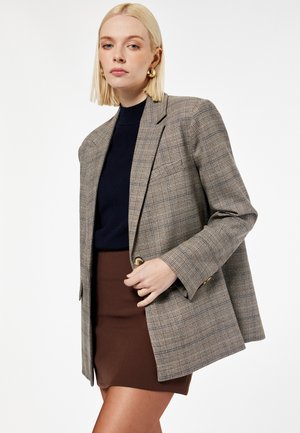 Lyshåret kvinde iført en beige ternet blazer over en sort top og brun nederdel, poserer foran en ensfarvet hvid baggrund.