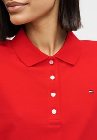 Γυναίκα που φοράει κόκκινο πόλο μπλουζάκι με τέσσερα κουμπιά, κοντά μαύρα μαλλιά και ένα μικρό ορθογώνιο λογότυπο στην δεξιά πλευρά του στήθους.