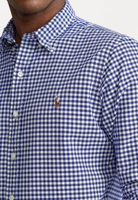 Camisa abotonada con patrón de cuadros vichy en azul y blanco. Cuenta con cuello, botones delanteros y un pequeño logotipo bordado en el pecho izquierdo.