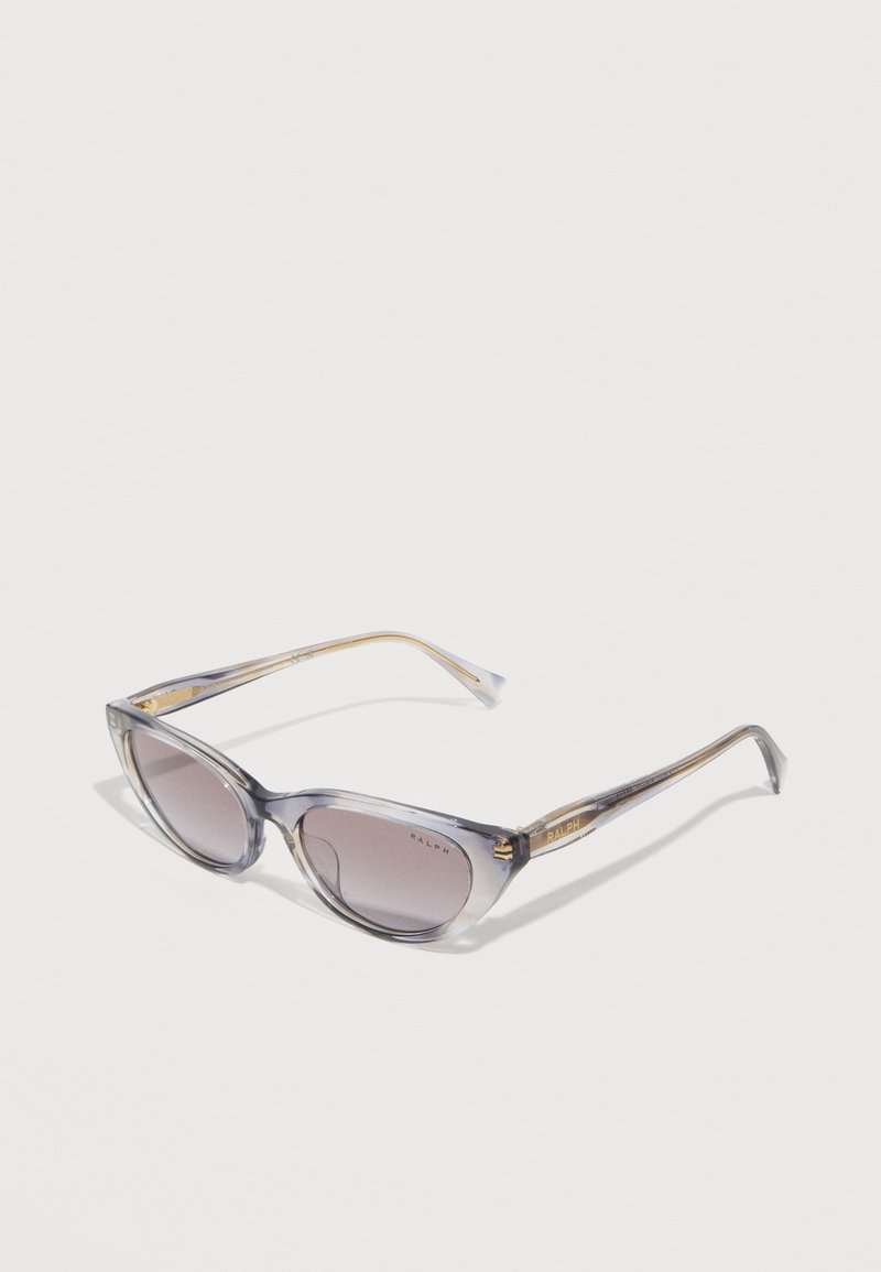 Διάφανες γκρι γυαλιά ηλίου σε σχήμα γάτας με φακούς με βαθμιαία απόχρωση και χρυσή σήμανση "Ralph" στα πλαϊνά, εμφανιζόμενα σε λευκό φόντο.