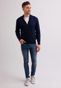 Granatowy kardigan z zamkiem błyskawicznym i wysokim kołnierzem, noszony na białej koszulce. Połączony z niebieskimi dżinsami i białymi trampkami. Gładka faktura materiału.