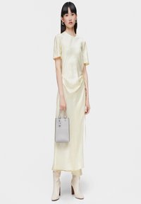 Abito di satin giallo con maniche corte, design drappeggiato e lacci laterali. Abbinato a una piccola borsa in pelle grigia con stampa "TOUS". Stivali beige con tacco.