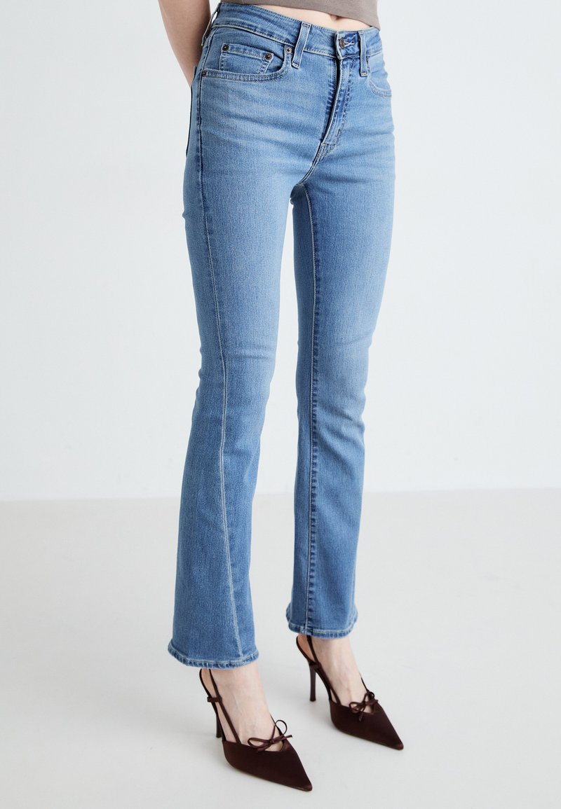 Jeans din denim albastru deschis, cu o croială slim și o ușoară evazare la tiv, având buzunare și o textură palidă. Asortați cu pantofi cu toc burgund.