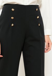 Visoke črne hlače z gladko teksturo, ki imajo štiri dekorativne zlate gumbe spredaj in prilagojen kroj.