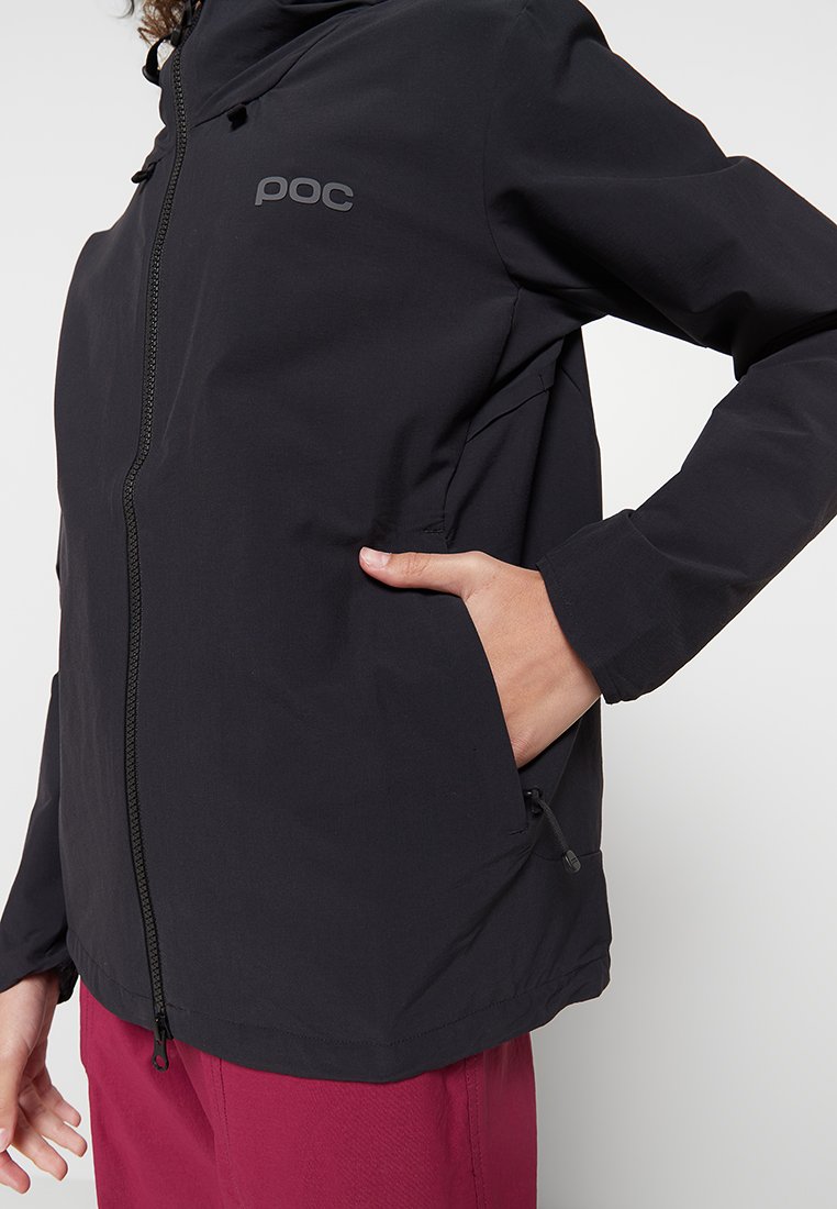 Persona vestida con chaqueta negra de cremallera y logo "poc", con la mano en el bolsillo lateral, y pantalones rosa oscuro contra un fondo simple.