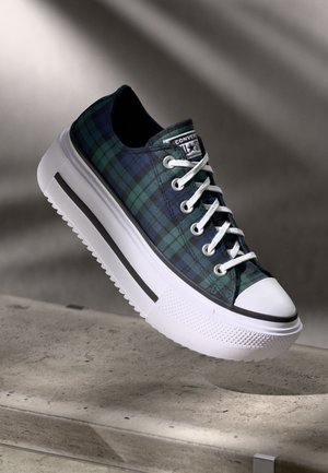 Alacsony szárú sneaker zöld és tengerészkék kockás anyagból, fehér vastag talppal, fehér fűzőkkel és fekete csíkkal a talpon, betonfelület felett bemutatva.