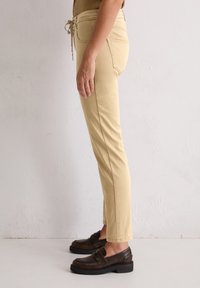 Persoon in beige slim-fit broek met een trekkoord in de taille en donkerbruine leren instappers, staande tegen een eenvoudige witte muur.