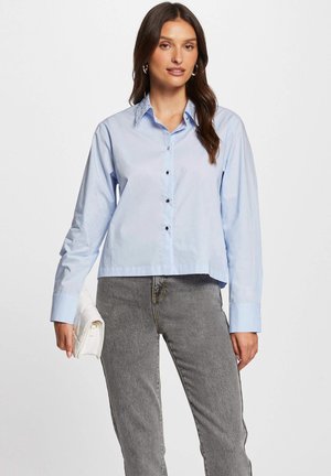 Lichtblauw overhemd met lange mouwen, voorzien van een puntige kraag en donkere knopen, gecombineerd met grijze jeans en een witte clutch.