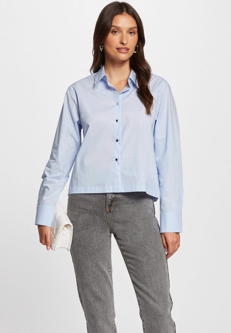 Lichtblauw overhemd met lange mouwen, voorzien van een puntige kraag en donkere knopen, gecombineerd met grijze jeans en een witte clutch.