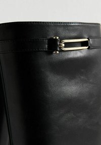 Bota alta de piel negra con textura suave. Cuenta con una cinta delgada y un detalle de hardware metálico en la parte superior para un ajuste seguro.