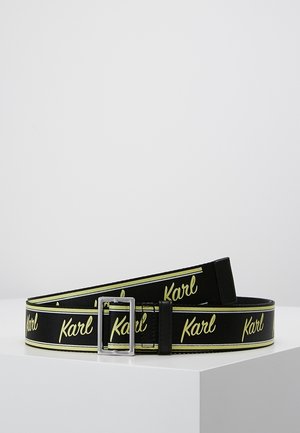 Musta vyö, jossa on toistuva keltainen "Karl"-kirjoitus, hopeanvärinen suorakaiteen muotoinen solki ja keltaiset raidalliset reunat, esillä valkoisella pinnalla vaaleaa taustaa vasten.