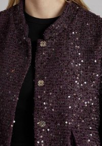 Primo piano di una persona che indossa una giacca di paillettes viola scuro sopra un top nero a coste, con bottoni rotondi metallici sulla giacca.