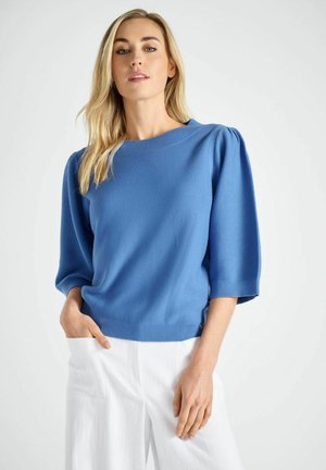 Blonde vrouw met een blauwe trui met pofmouwen en witte broek, staand met één hand in de broekzak tegen een effen achtergrond.