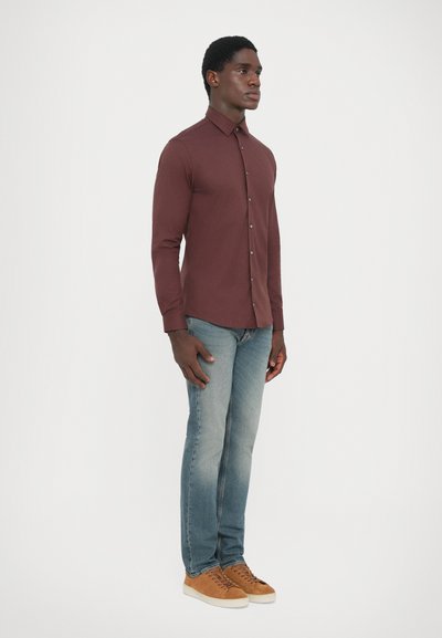 Camisa de botón color burdeos con mangas largas, combinada con jeans de un azul claro y zapatillas marrones. Diseño simple, textura suave, con detalles mínimos.