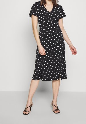 Femme portant une robe midi noire à pois blancs, manches courtes, encolure en V et fentes latérales, associée à des sandales à talons noires à brides.
