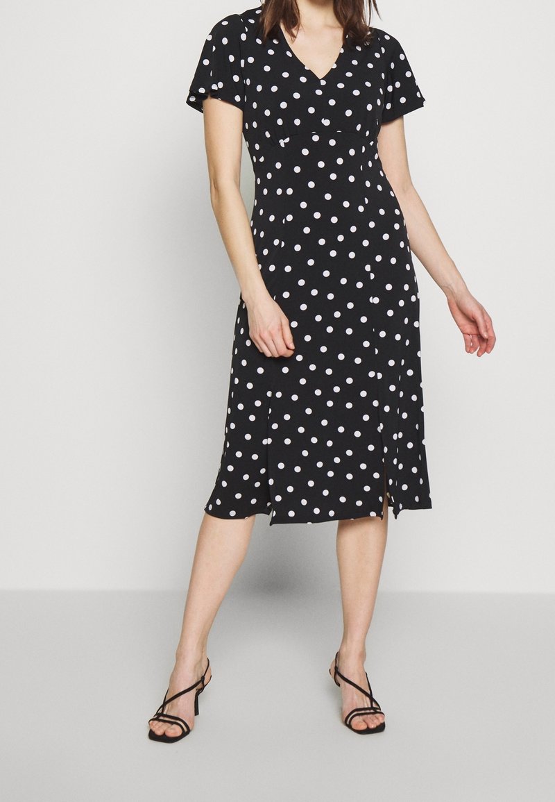 Femme portant une robe midi noire à pois blancs, manches courtes, encolure en V et fentes latérales, associée à des sandales à talons noires à brides.