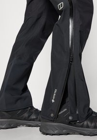 Crne vodootporne hlače s GORE-TEX oznakom, s punim zatvaračem i blago suženim dizajnom, uparene s crnim obućom za planinarenje s teksturom.