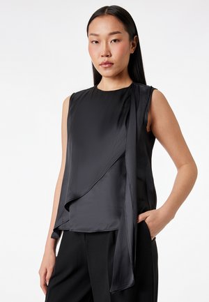 Kvinde med langt, mørkt hår iført ærmeløs, sort lagdelt bluse og sorte bukser, stående med den ene hånd i lommen mod en hvid baggrund.