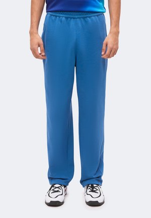 Mand iført løstsiddende blå joggingbukser og hvide sneakers med sorte snørebånd, stående mod en enkel baggrund.