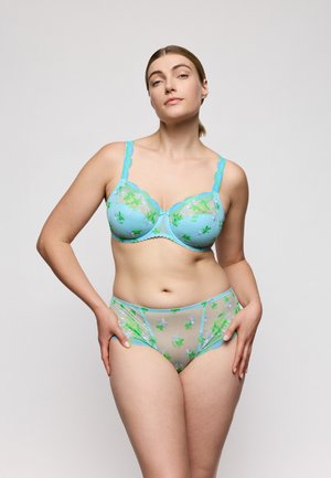 Vrouw draagt doorschijnend lichtblauw lingerie met groene bloemenborduursels, staand met handen op de heupen tegen een effen achtergrond.