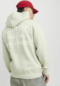 Hellgrüner Hoodie mit großem geprägtem Text auf dem Rücken; hat eine Kapuze mit Kordelzug und gerippte Bündchen. Kombiniert mit grauen Jeans.