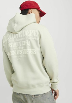Light green hoodie with large embossed text on the back; features a drawstring hood and ribbed cuffs. Paired with gray denim.