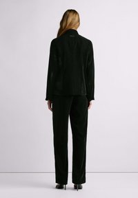 Inimene pikkade juustega, kes kannab musta sametkostüümi ja musti kõrge kontsaga kingi, seistes seljaga vaataja poole tavalise hele tausta ees.