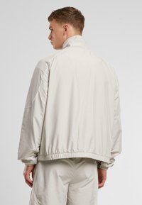 Beige, lett jakke med høy krage, elastisk liv og mansjetter. Har kontrasterende hvite sømmer langs skuldrene.