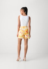 Top senza maniche bianco a coste abbinato a pantaloni corti floreali gialli con motivi rosa e arancioni; la modella indossa sandali con tacco chiari.